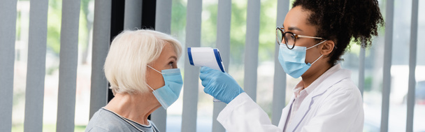 African american doctor in medical mask holding pyrometer near senior patient, banner  - Photo, Image