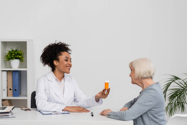 Sorridente medico afro-americano con in mano un barattolo con pillole vicino al paziente in ospedale  - Foto, immagini