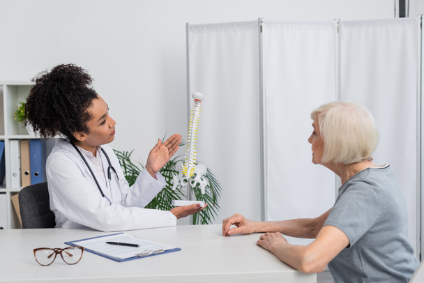 Fisioterapeuta afroamericano apuntando al modelo de columna vertebral mientras habla con un paciente mayor  - Foto, imagen