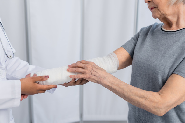 Ausgeschnittene Ansicht einer Seniorin mit Gipsverband am Arm, die in der Nähe eines amerikanischen Arztes steht  - Foto, Bild
