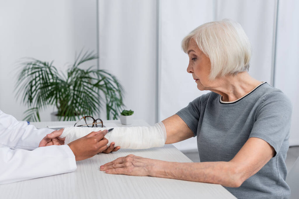 Vista lateral del médico afroamericano que señala el vendaje de yeso en el brazo de una mujer anciana en la clínica  - Foto, imagen