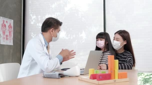 Una madre con una máscara facial lleva a su hija enferma a una cita de examen con un médico asiático en el departamento de pediatría de una clínica infantil. Tratar enfermedades y consultar sobre salud familiar. - Metraje, vídeo