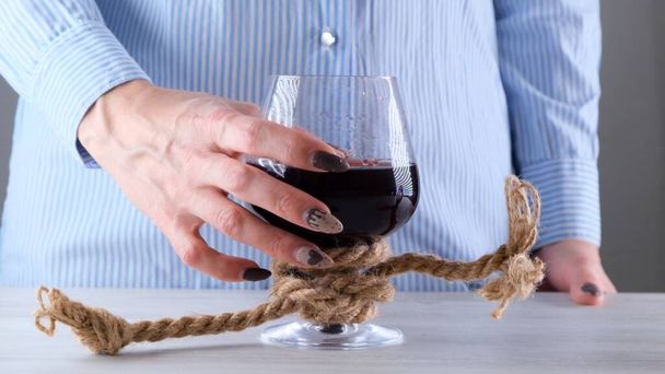 θηλυκό χέρι κρατώντας ποτήρι κρασί είναι δεμένο με ένα σχοινί γιούτα. Η έννοια της εξάρτησης από το αλκοόλ. - Φωτογραφία, εικόνα