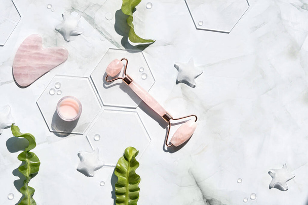 Pink Quarts stone face roller, cream, hexagons. Gua sha stone for beauty facial massage therapy. Natural cosmetics laboratory. Off white flat lay with palm leaf shadow, copy-space. - Foto, imagen