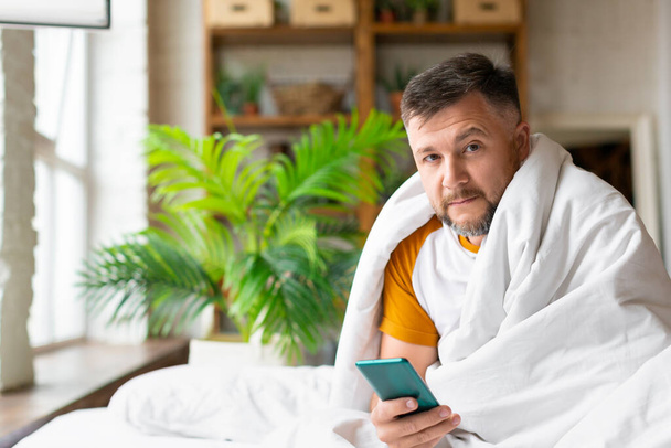 病気の中年の男が毛布の下のベッドの上に座っている。彼はスマートフォンを持っており、オンライン相談のために彼の医者を呼び出すつもりです。. - 写真・画像