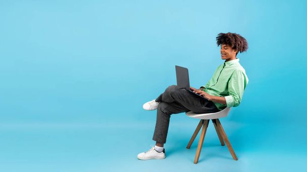 Eラーニングの概念。アフリカ系アメリカ人の学生のラップトップを使用して、青い背景、パノラマ、フリースペースに椅子に座って - 写真・画像