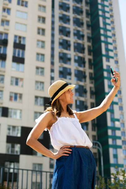 Young beautiful woman blogger in summer clothing making selfie in front of modern building - Photo, Image