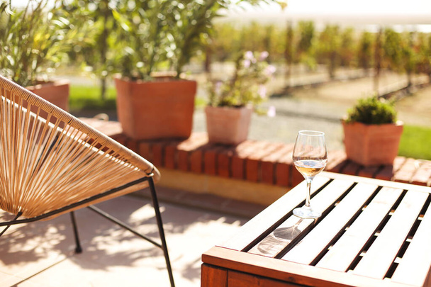 Glass of white wine on rustic wooden table and flower pots on terrace outside with vineyard hills landscape on sunny day morning in summer time - Photo, Image