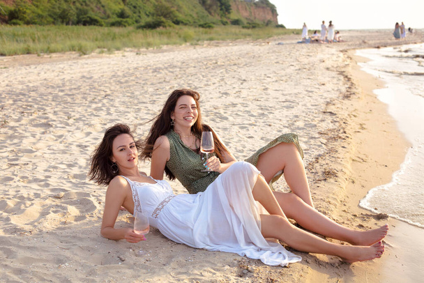 Cieszę się, że dorosłe koleżanki w letnim stroju uśmiechają się, ciesząc się winem na piaszczystej plaży w pobliżu morza - Zdjęcie, obraz