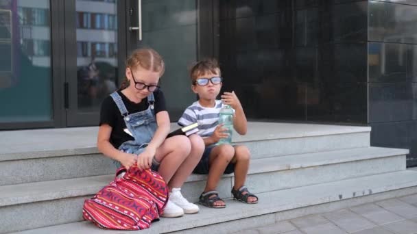 Dos niños en gafas con libros y mochilas almorzan - Imágenes, Vídeo