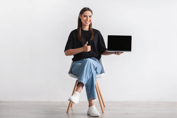 Sandalyeye oturmuş gülümseyen genç bir kadın, boş ekranlı dizüstü bilgisayarını gösteriyor, işaret ediyor, web sitesi için maket yapıyor. - Fotoğraf, Görsel