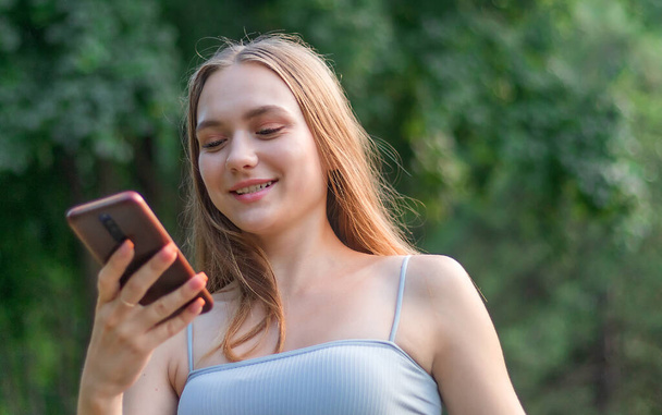 mladá charismatická krásná evropská dívka s dlouhými blond vlasy drží telefon v rukou a usmívá se, ženský portrét dospívající dívky - Fotografie, Obrázek