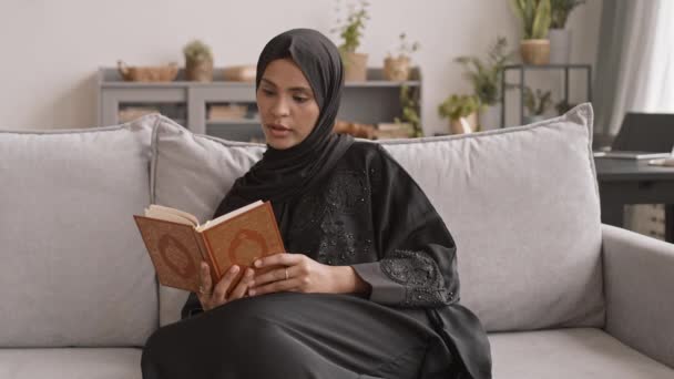 PAN μέσο αργό πλάνο του νεαρή μουσουλμάνα γυναίκα με παραδοσιακά μαύρα ρούχα και μαντίλα ανάγνωση Κοράνι δυνατά, ενώ κάθεται στον καναπέ στο σαλόνι - Πλάνα, βίντεο