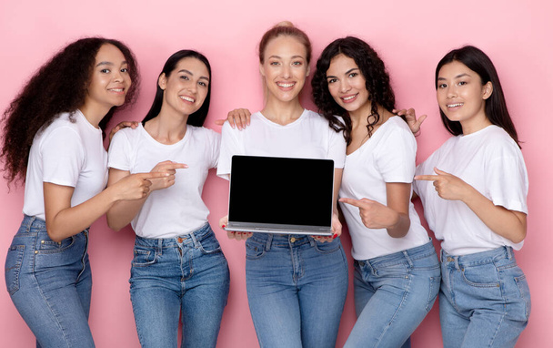 Diversas mujeres sosteniendo el ordenador portátil apuntando a la pantalla vacía, fondo rosa - Foto, Imagen