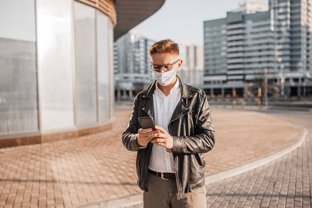 Bonito homem com uma máscara protetora em seu rosto com óculos com um smartphone na rua de uma cidade grande. Empresário falando ao telefone no fundo urbano - Foto, Imagem