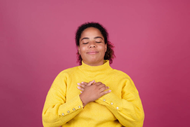 Piękna Afroamerykanka na różowym tle szczęśliwe uśmiechnięte ręce na klatce piersiowej miłość się koncepcja, self care - Zdjęcie, obraz