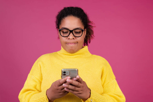 Bella donna afroamericana su sfondo rosa digitando messaggio sul telefono cellulare concentrato premuroso - Foto, immagini