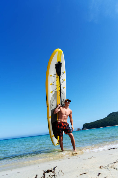 A man stands on a sandy shore and holds a large yellow paddle board. - 写真・画像
