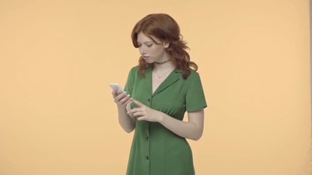 redhead teenage girl smiling while looking at smartphone isolated on yellow - Footage, Video