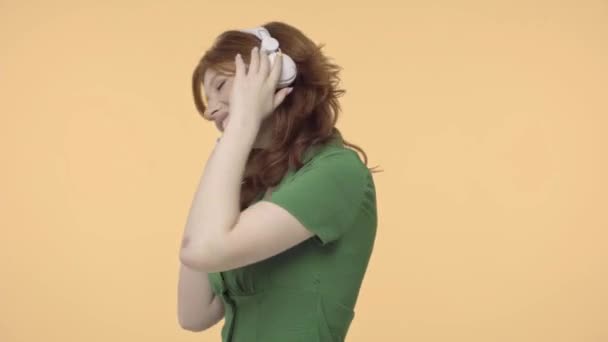 Ευτυχισμένη κοκκινομάλλα έφηβη με ακουστικά που χορεύει απομονωμένη στο κίτρινο - Πλάνα, βίντεο