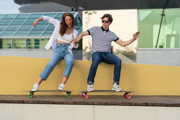 Jeune couple hipster s'amuser avec longboard prétendre monter skateboard assis dans skatepark urbain - Photo, image
