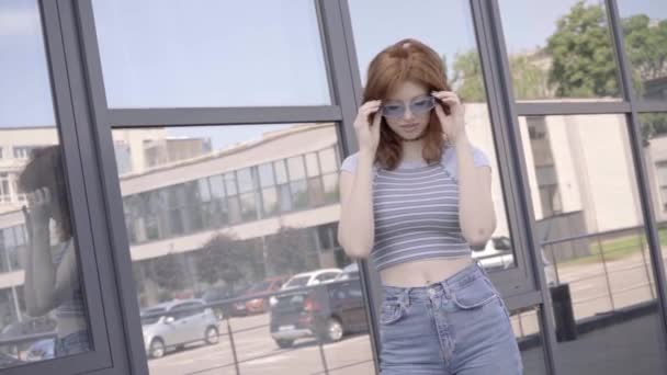 glückliches rothaariges Teenager-Mädchen nimmt Sonnenbrille in der Nähe von Gebäude ab  - Filmmaterial, Video