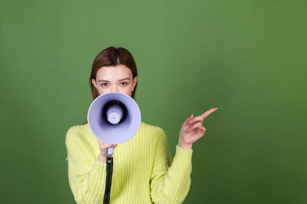 Young woman with perfect natural makeup brown big lips in casual sweater on green background with megaphone  happy positive  point finger right - Photo, Image