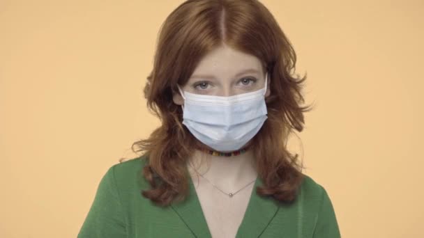 рыжая девушка-подросток в медицинской маске смотрит на камеру, изолированную на желтом - Кадры, видео