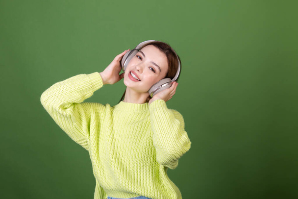 Young woman with perfect natural makeup brown big lips in casual sweater on green background with wireless headphones happy  dancing moving around - Foto, afbeelding