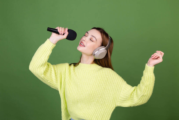 Mujer joven con maquillaje natural perfecto labios grandes marrones en suéter casual sobre fondo verde con auriculares inalámbricos sostienen el micrófono cantando - Foto, imagen