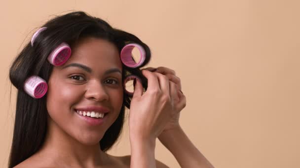 afro-americano mulher curling cabelo aplicando curlers no fundo bege - Filmagem, Vídeo