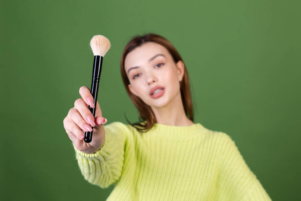 Jonge vrouw met perfecte natuurlijke make-up bruine grote lippen in casual trui op groene achtergrond houden blush borstel poseren - Foto, afbeelding
