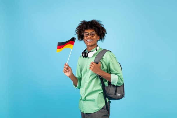 Modern eğitim ve bilgi alışverişi. Sırt çantası ve küçük Almanya bayrağı olan, mavi arka planlı Afrikalı Amerikalı genç adam. - Fotoğraf, Görsel