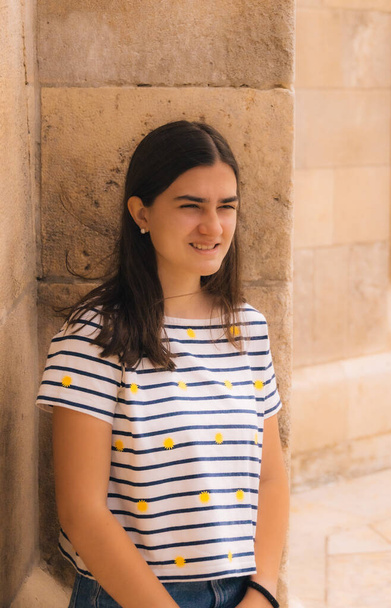 portrait of a young teenage girl with long hair smiling in a striped t-shirt with yellow suns against the background of a stone wall on a sunny day - Foto, Imagem