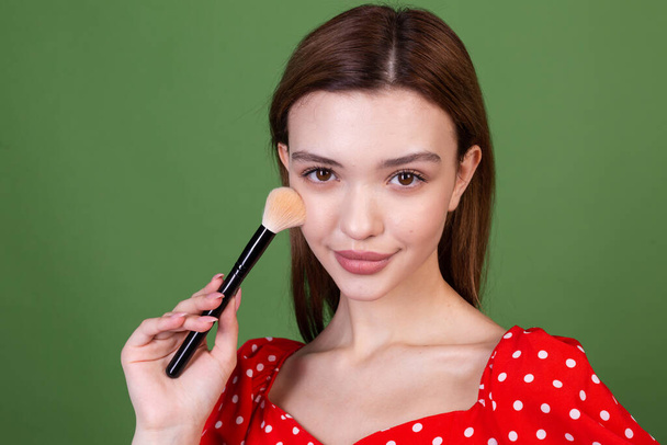 Young woman with perfect natural makeup brown big lips in polka dot red dress on green background holds blush brush fashion beauty portrait - Photo, Image