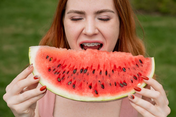 Close-up portrait of red-haired young woman with braces eating watermelon outdoors - Fotografie, Obrázek
