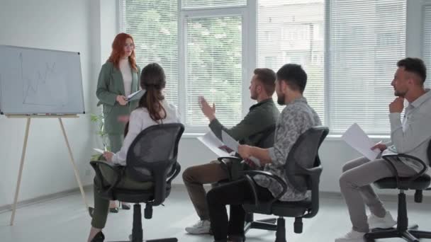 mladá podnikatelka prezentuje nový projekt na flipchartu pro ženy a muže, mladý muž zvedne ruce a ptá se na otázky během obchodní schůzky v kanceláři - Záběry, video