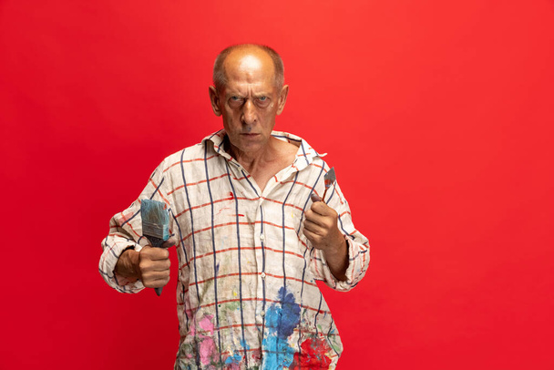 Pintor gris maduro o senior con un pincel delante de la cámara. Hombre divertido en una camisa manchada de pintura tonteando alrededor aislado en rojo - Foto, Imagen