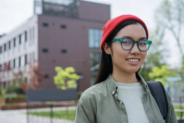 Piękny uśmiechnięty azjatycki student w okularach spacerujący po kampusie uniwersyteckim. Wracając do szkoły, koncepcja edukacji. Szczęśliwa kobieta turystka z plecakiem patrząc na kamerę stojącą na ulicy - Zdjęcie, obraz