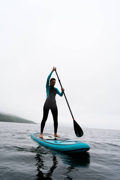 A woman in a wetsuit paddle a paddleboard with an oar on the sea waves in cloudy weather. - Фото, зображення