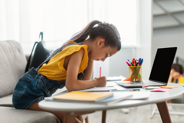 Egy lány ül az asztalnál, üres laptoppal, tankönyvbe írva. - Fotó, kép