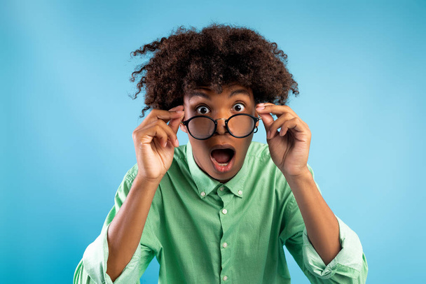 émotionnel afro-américain adolescent guy dans lunettes ouverture bouche dans surprise, en regardant la caméra sur fond bleu - Photo, image