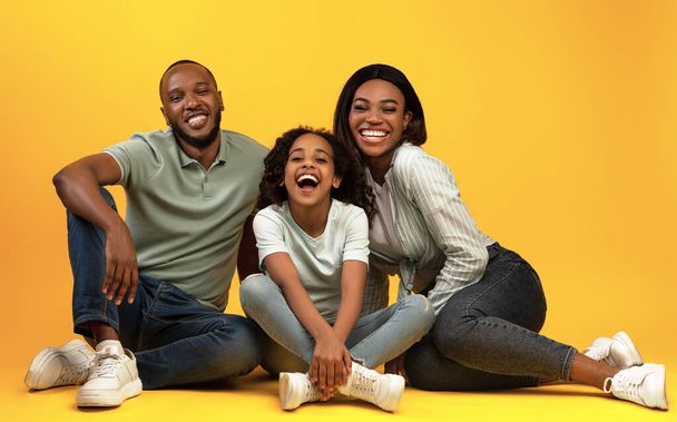 Felice famiglia afro-americana seduta sul pavimento e ridere, sorridendo insieme alla fotocamera su sfondo giallo studio - Foto, immagini