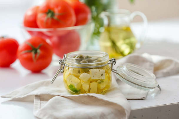 Lasipurkki maukasta fetajuustoa, öljyä, oliiveja ja tomaattia kevyellä pöydällä, lähikuva - Valokuva, kuva