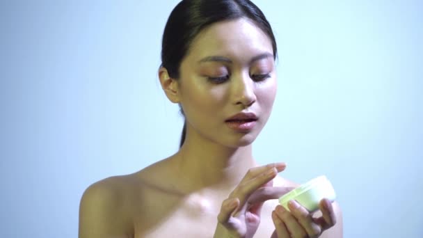 bonita mujer asiática aplicando crema sobre suave piel sobre azul - Metraje, vídeo