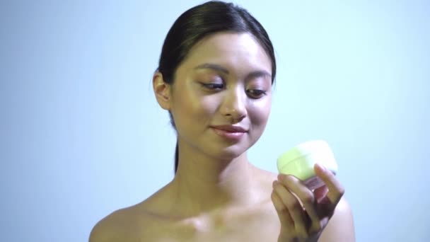 feliz joven asiático mujer celebración contenedor con crema en azul - Imágenes, Vídeo