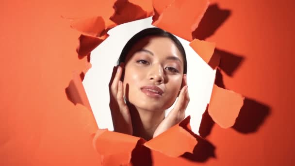 joven asiático mujer tocando perfecto piel cerca rasgado papel naranja fondo - Imágenes, Vídeo