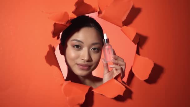felice donna asiatica bottiglia con siero vicino strappato sfondo di carta arancione - Filmati, video