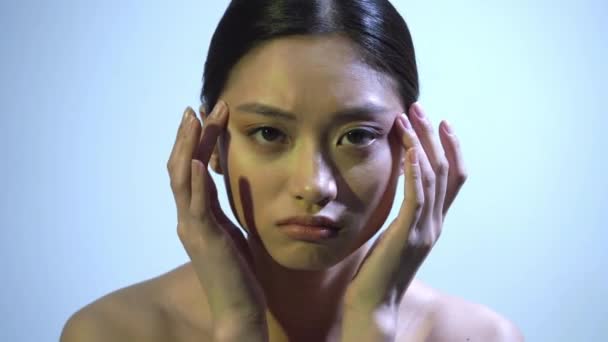 trieste jonge aziatische vrouw met blote schouders aanraken gezicht op blauw - Video