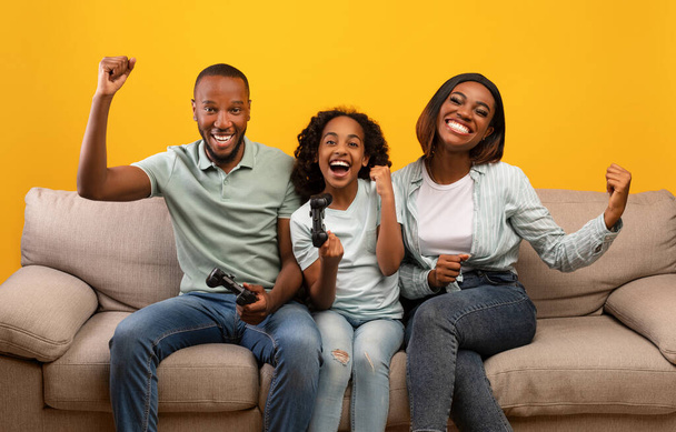 Actividades familiares. feliz negro padres y hija jugando video juegos, sentado en sofá sobre amarillo fondo - Foto, Imagen
